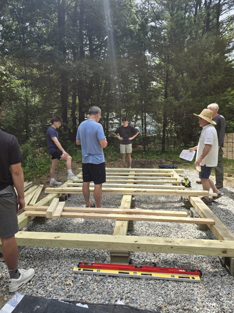 Volunteers standing around the pressure-treated floor frame set on concrete blocks over the finished gravel pad, one holding the plans