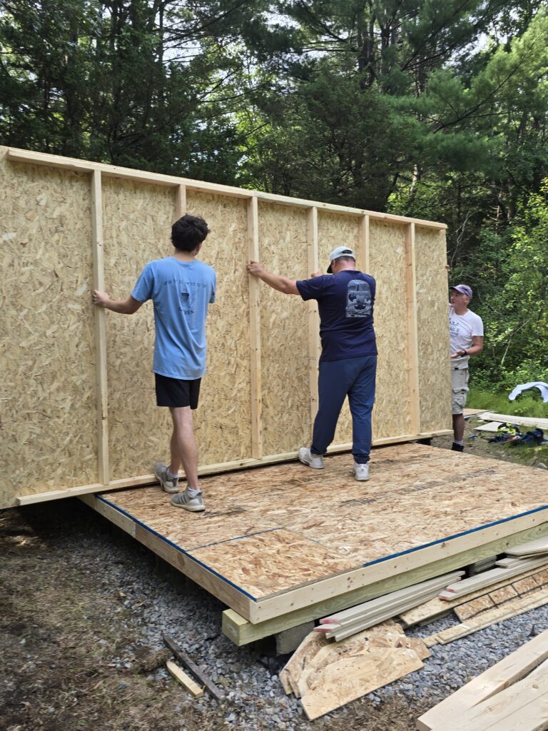 Three people tilting a framed and sheathed wall upright onto the shed's plywood deck