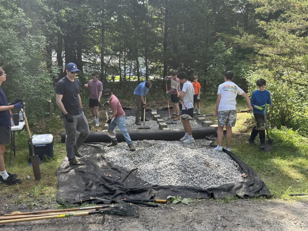 More than a dozen volunteers of all ages raking and shovelling a large pile of crushed stone spread over landscape fabric