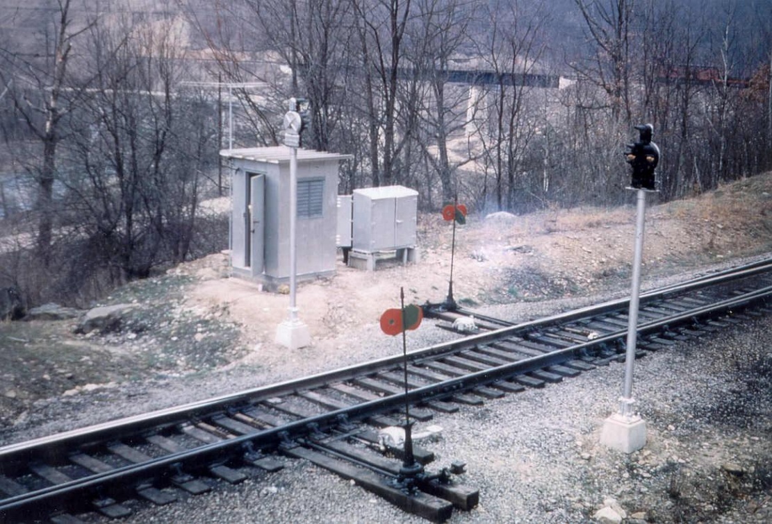 Grey metal equipment cases beside a railroad switch, with signal masts and switch targets
