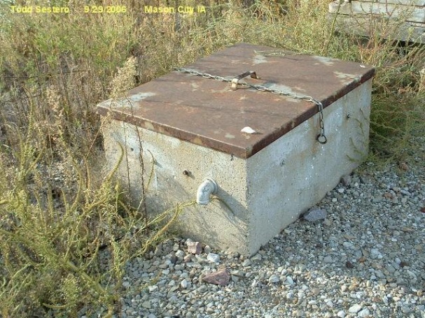 A rectangular concrete battery box with a rusted steel lid secured by a chain, standing in gravel beside a track