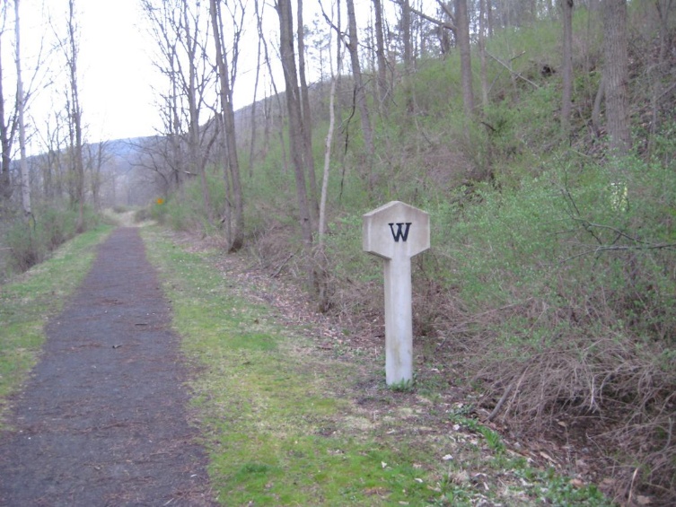 A whistle post marked with the letter W standing beside a gravel trail through bare spring woods